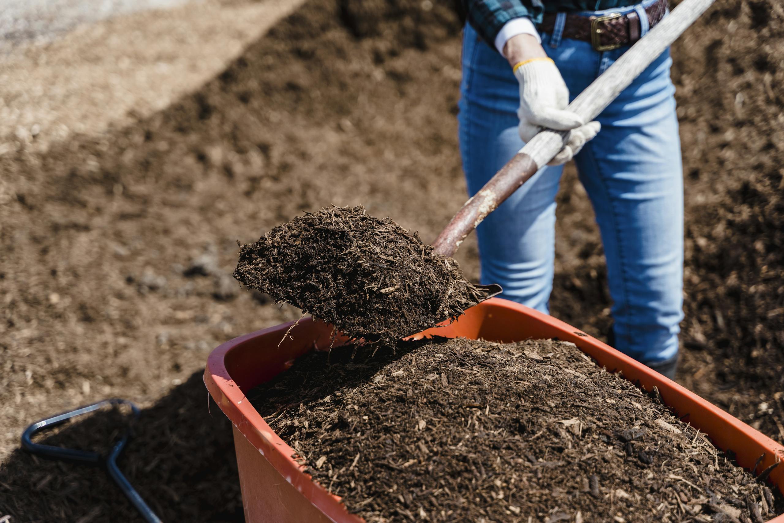 A person shovels compost into a red container, preparing soil for gardening.